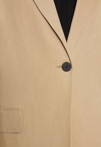 Blazer beige con solapa en pico, tela texturizada, cierre de un botón negro y bolsillo de parche en el lado izquierdo.