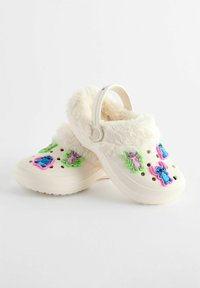 Sabots en peluche blancs avec des charms de personnages colorés. Forme arrondie, trous de ventilation et doublure en fourrure douce autour du col.