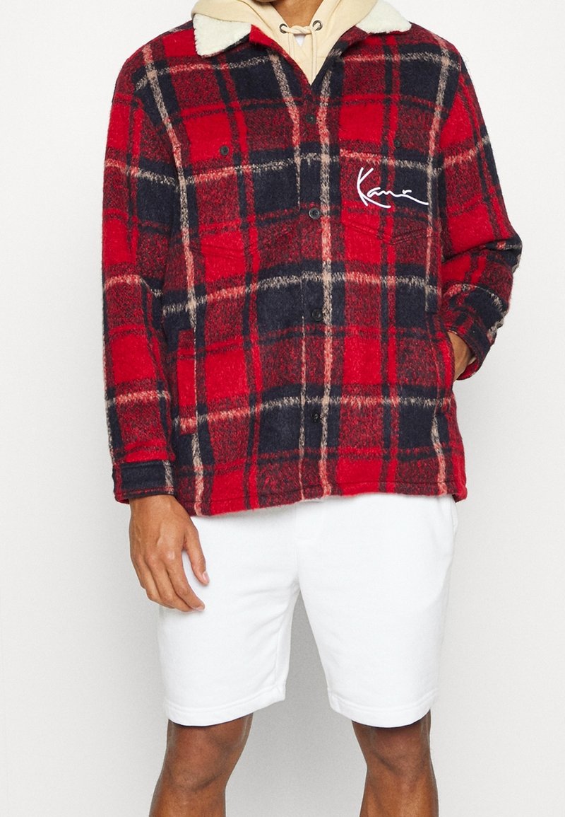Homme portant une veste à carreaux rouge et bleu marine avec col en peau lainée blanche sur un sweat à capuche beige et un short blanc, mains détendues sur les côtés.