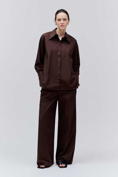 Femme les mains dans les poches, vêtue d'une chemise ample marron foncé et d'un pantalon large, debout devant un fond gris clair uni.