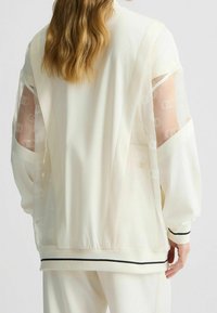 Bílá lehká bunda s průhlednými síťovými rukávy. Má jemný vzor loga, žebrované manžety a černý akcent na spodním lemu.