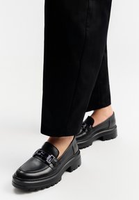 Czarne skórzane mokasyny z masywną podeszwą, z dekoracyjnym metalowym łańcuchem z przodu. Gładka tekstura, łatwe do zakładania.
