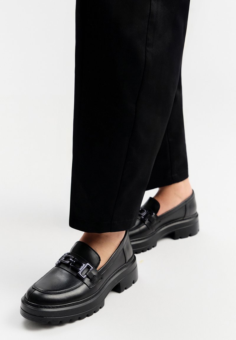 Czarne skórzane mokasyny z masywną podeszwą, z dekoracyjnym metalowym łańcuchem z przodu. Gładka tekstura, łatwe do zakładania.