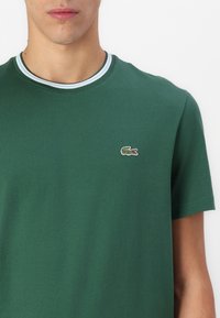Tricou din bumbac verde, cu decolteu tip rundă și margine în dungi albastre și albe, având un mic logo brodat cu crocodil pe piept.