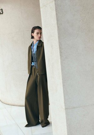 Olive green tailored jacket and wide-leg pants paired with a blue shirt and a gray vest, with smooth textures in a minimalist setting.