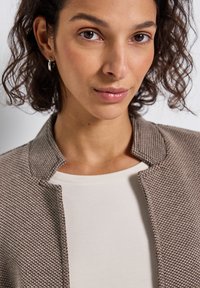 Veste marron texturée avec un petit col, portée sur un haut crème. Présente un gros plan du visage du modèle et de ses cheveux bouclés.