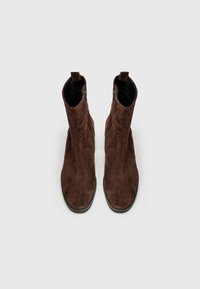 Botas de tobillo de ante marrón con una textura suave, cremalleras laterales y un diseño de puntera redondeada. Suelas de goma planas y detalles de costura sutiles.
