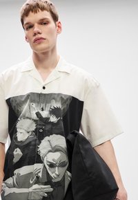 Νέος άνδρας φοράει λευκό κοντομάνικο πουκάμισο με μαυρόασπρες εκτυπώσεις χαρακτήρων anime, κρατώντας μια μαύρη ζακέτα στο δεξί του χέρι.