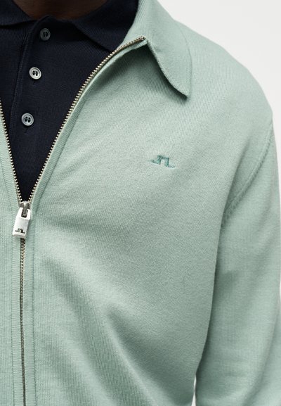Άνδρας που φοράει ανοιχτό πράσινο πουλόβερ με φερμουάρ πάνω από μπλε σκουρόχρωμο πόλο με κουμπιά, εμφανίζοντας το φερμουάρ και μικρό κεντημένο λογότυπο στο στήθος.