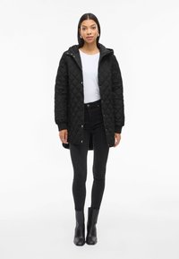 Črna prešita jakna s kapuco, saponasti zap closures in rebrastimi manšetami, v kombinaciji z belo majico, črnimi kavbojkami in gležnjarji.