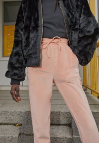 Černá umělá kožešinová bomber bunda přes černý top; růžové sametové tepláky se šněrováním v pase a bílým logem. Hladká textura, volný střih.