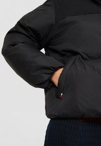 Chaqueta acolchada negra con cuello alto, puños elásticos y un bolsillo lateral. Presenta una pequeña etiqueta con el logo en rojo, blanco y azul en el bolsillo.