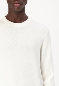 Maglione bianco a costine con collo rotondo, caratterizzato da un sottile logo ricamato. Il tessuto sembra morbido con una finitura texturizzata. Design minimalista.
