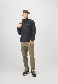 Marine fleece pullover med lynlås ved halsen, parret med olivengrønne cargo bukser. Modellen bærer en grøn cap og sorte vandresko med lyse accenter.