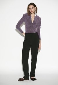 Lila Langarmtop mit V-Ausschnitt und strukturiertem Stoff, kombiniert mit schwarzen, taillierten Hosen und verzierten schwarzen Loafers.