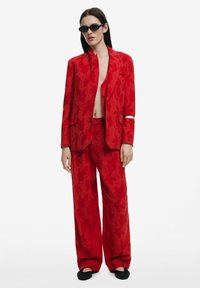 Rood bloemenpak met een getailleerde blazer en brede broek met een subtiele glans. Accessoires zijn onder andere donkergekleurde zonnebrillen en zwarte schoenen.