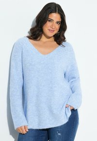 Lächelnde Frau mit dunklen Haaren, die einen hellblauen V-Ausschnitt-Pullover und Jeans trägt und mit einer Hand in der Tasche vor einem weißen Hintergrund posiert.