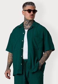 Ein tätowierter Mann in einem türkisfarbenen Kurzarmhemd und passender Hose, der Sonnenbrillen trägt, steht selbstbewusst vor einem neutralen Hintergrund.