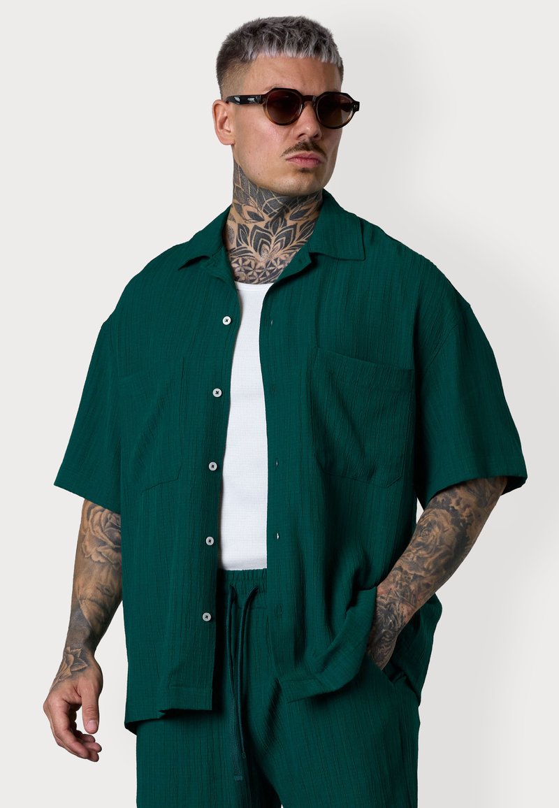 Ein tätowierter Mann in einem türkisfarbenen Kurzarmhemd und passender Hose, der Sonnenbrillen trägt, steht selbstbewusst vor einem neutralen Hintergrund.