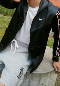 Svart zip-up jacka med huva, grå shorts med en svart sol- och regnbågslogotyp, vitt t-shirt under, och dragsko-shorts.