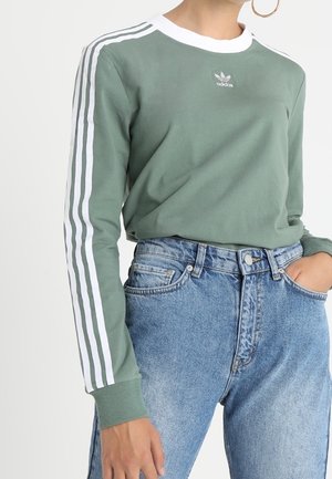 Kobieta w zielonej bluzie Adidas z białymi paskami na rękawach oraz wysokim stanem niebieskich dżinsach, z ręką w kieszeni i widoczną dużą kolczykiem kołowym.