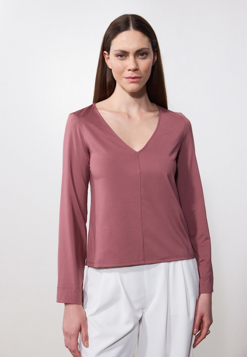 Top de manga larga y cuello en V en color rosa suave, hecho de una tela lisa con un detalle de costura mínima, combinado con pantalones blancos.