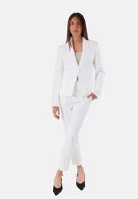 Blazer e pantaloni bianchi su misura realizzati in tessuto liscio. Il blazer presenta un singolo bottone e rever a punta, abbinato a tacchi neri.