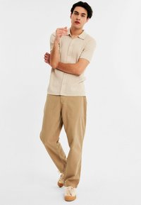 Beige ribbed short-sleeve polo shirt with a button-up front, paired with light brown pants and light sneakers, standing against a white background.