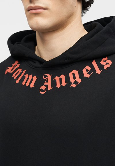 Jaunas žmogus dėvi juodą gobtuvą su raudonu gotikinio stiliaus užrašu „Palm Angels“ per krūtinę ir gobtuvą.