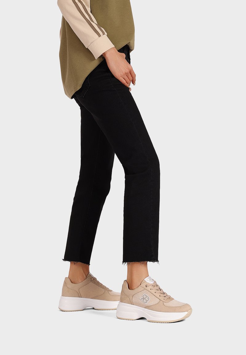 Sneaker beige con una parte superiore in suede e mesh lisci, caratterizzati da una suola bianca spessa e dettagli con logo. Abbinati a jeans neri a vita bassa.