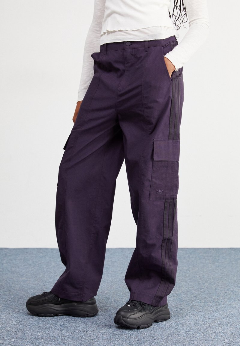 Fialové cargo kalhoty se bočními kapsami, s černými pruhy po stranách, vyrobené z odolné tkaniny. Nosené s černými teniskami.