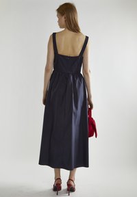 Marineblauwe, mouwloze jurk met een aansluitend lijfje en een geplooid rokje. Ritssluiting aan de achterkant en een effen kleur, gecombineerd met een rode handtas en hakken.