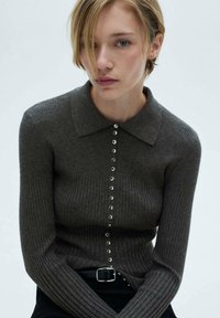 Ribbet, mørkegrå strikcardigan med en foldet krave og en knaplukning fortil, der har glinsende sølvknapper. Lange ærmer, tætsiddende facon.