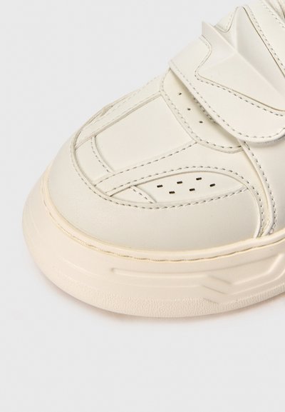Μπεζ δερμάτινο αθλητικό παπούτσι με διάτρητες λεπτομέρειες, ορατή ραφή και παχύ, υφημένο πέλμα σε λευκό φόντο.