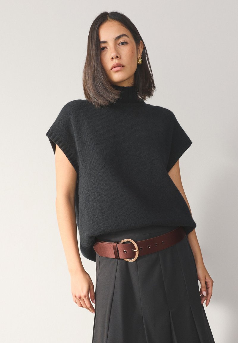 Femme aux cheveux châtains mi-longs portant un pull sans manches noir à col roulé, une large ceinture marron et un pantalon plissé foncé.