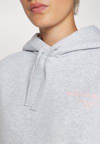 Felpa grigia chiara con cappuccio e lacci, caratterizzata da una texture morbida e un dettaglio del logo rosa che recita "BJÖRN BORG STOCKHOLM 1956."