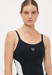 Donna con capelli tirati indietro che indossa un costume da bagno Arena bianco e nero, rivolta in avanti con un'espressione neutra su uno sfondo semplice.