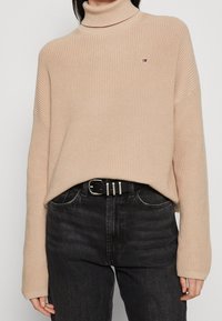 Beżowy sweter z golfem w prążki w połączeniu z czarnymi dżinsami i czarnym skórzanym paskiem z srebrną klamrą.