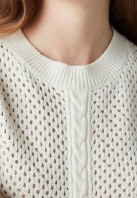 Primo piano del collo e delle spalle di una donna che indossa un maglione a maglia color crema con un motivo a trecce e un design a rete aperta.