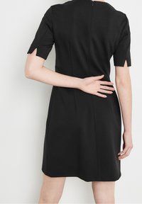 Robe noire à manches courtes, avec un décolleté rond, une fermeture éclair dans le dos et des fentes au bas des manches, fabriquée en tissu lisse avec une silhouette cintrée.