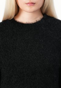 Nærbilde av en person med rett blondt hår som har på seg en teksturert svart, lodden genser, viser nakke, lepper og del av ansiktet.