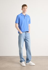 Hellblaues Baumwoll-Poloshirt mit kurzen Ärmeln, kombiniert mit hellblauen Jeans; das Modell trägt lässig weiße Sneakers.