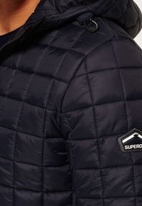 Laivastonsininen toppatakki tikatulla tekstuurilla, jossa on hupullinen ja logoplaatti. Kangas näyttää kiiltävältä ja kevyeltä.