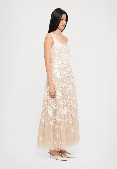 Femeie în picioare, cu fața întoarsă lateral, îmbrăcată într-o rochie maxi fără mâneci, bej, cu dantelă florală, și cu tocuri kitten crem, pe un fundal alb.