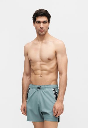 Młody mężczyzna z ciemnymi włosami, ubrany w turkusowe kąpielówki z czarnymi i białymi bocznymi paskami, stoi na jednolitym jasnym tle.