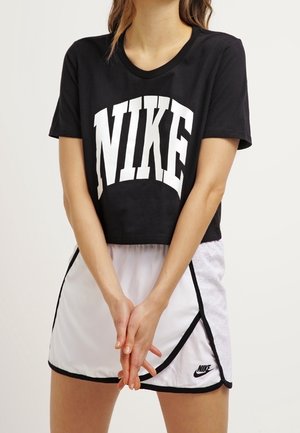 Kobieta w czarnej, skróconej koszulce Nike i białych sportowych szortach Nike z czarną lamówką, stojąca z rękami splecionymi z przodu.