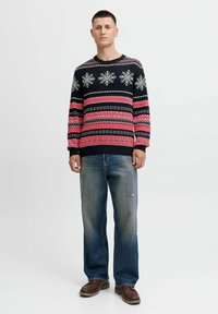Maglione lavorato a maglia in navy con motivi di fiocchi di neve bianchi e geometrici, accenti rossi, collo rotondo, abbinato a jeans blu sbiaditi e scarpe marroni.