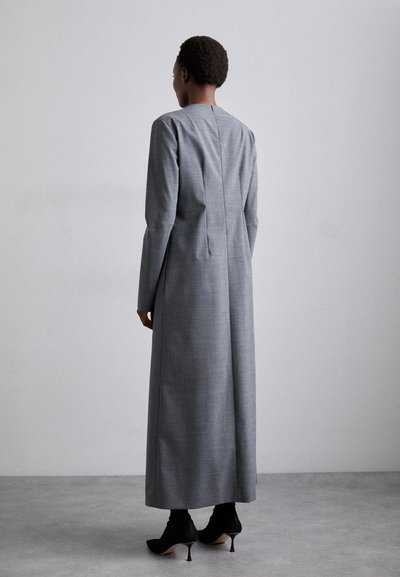 Larga vestido gris hecho de una tela texturizada, con mangas largas y una abertura en la parte trasera. Llevado con tacones negros puntiagudos.