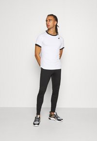 Weißes Sport-T-Shirt mit schwarzem Kontrastbesatz, kombiniert mit schwarzen Leggings und schwarzen Sneakers mit goldenen Akzenten.