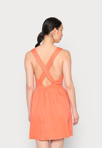 Vestido sem mangas cor-de-coral com alças cruzadas nas costas, cintura ajustada e design de costas abertas. Feito de um tecido leve, até ao joelho.